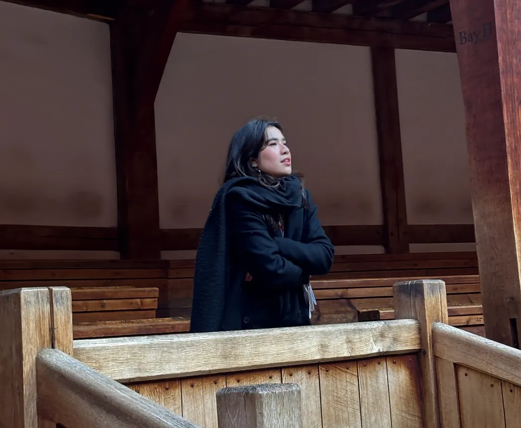 Woman in a black coat and scarf stands on a wooden balcony, arms crossed, looking off to the side inside a traditional wooden structure.