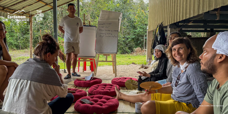 Instructor leads an outdoor workshop under a wooden shelter, barefoot participants sit on red cushions around a flip chart. Forest backdrop with logos in corner.